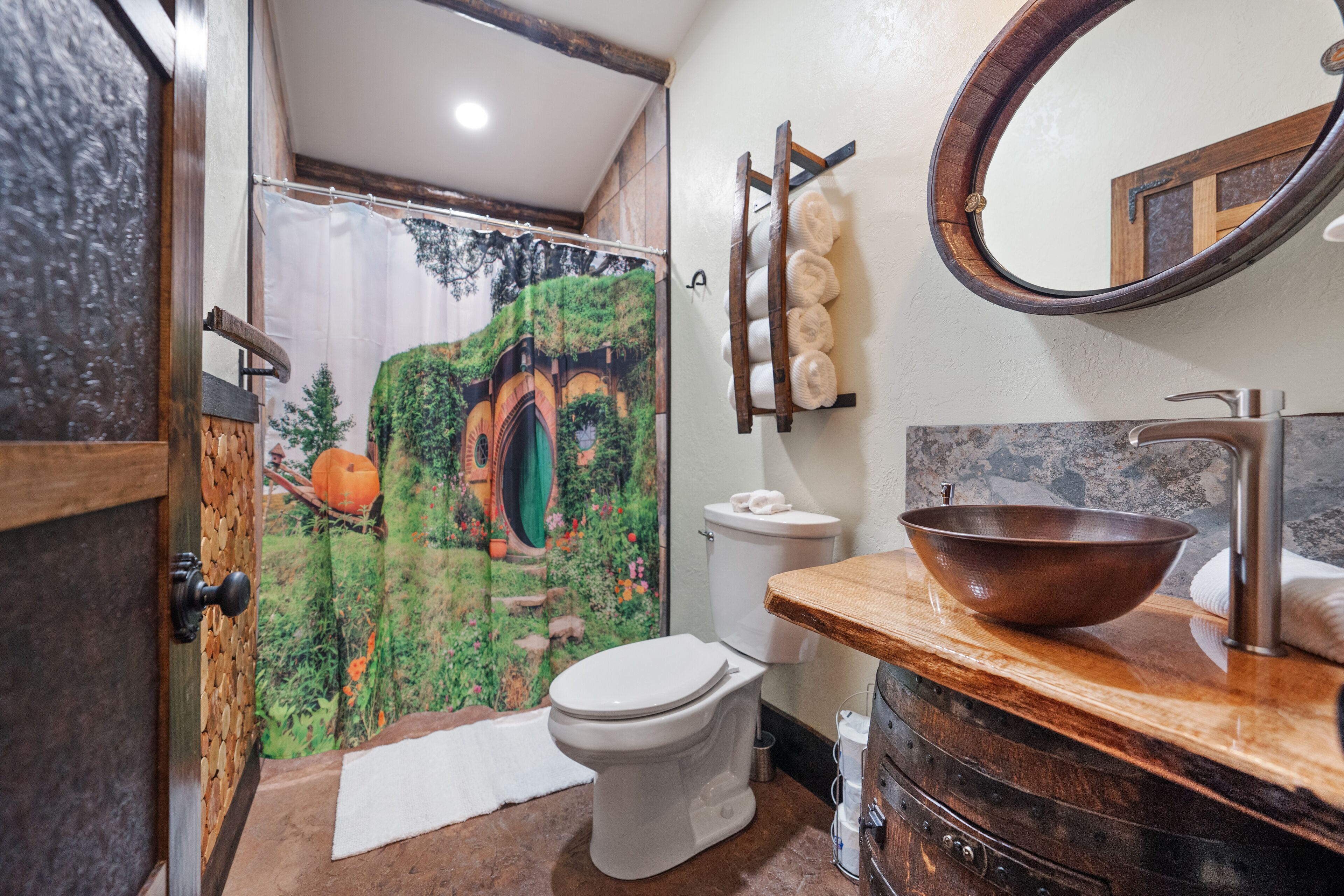 A bathroom with a rustic design, featuring a wooden door, a shower curtain with a forest scene, a white toilet, a wooden vanity with a copper basin, and a mirror on the wall.