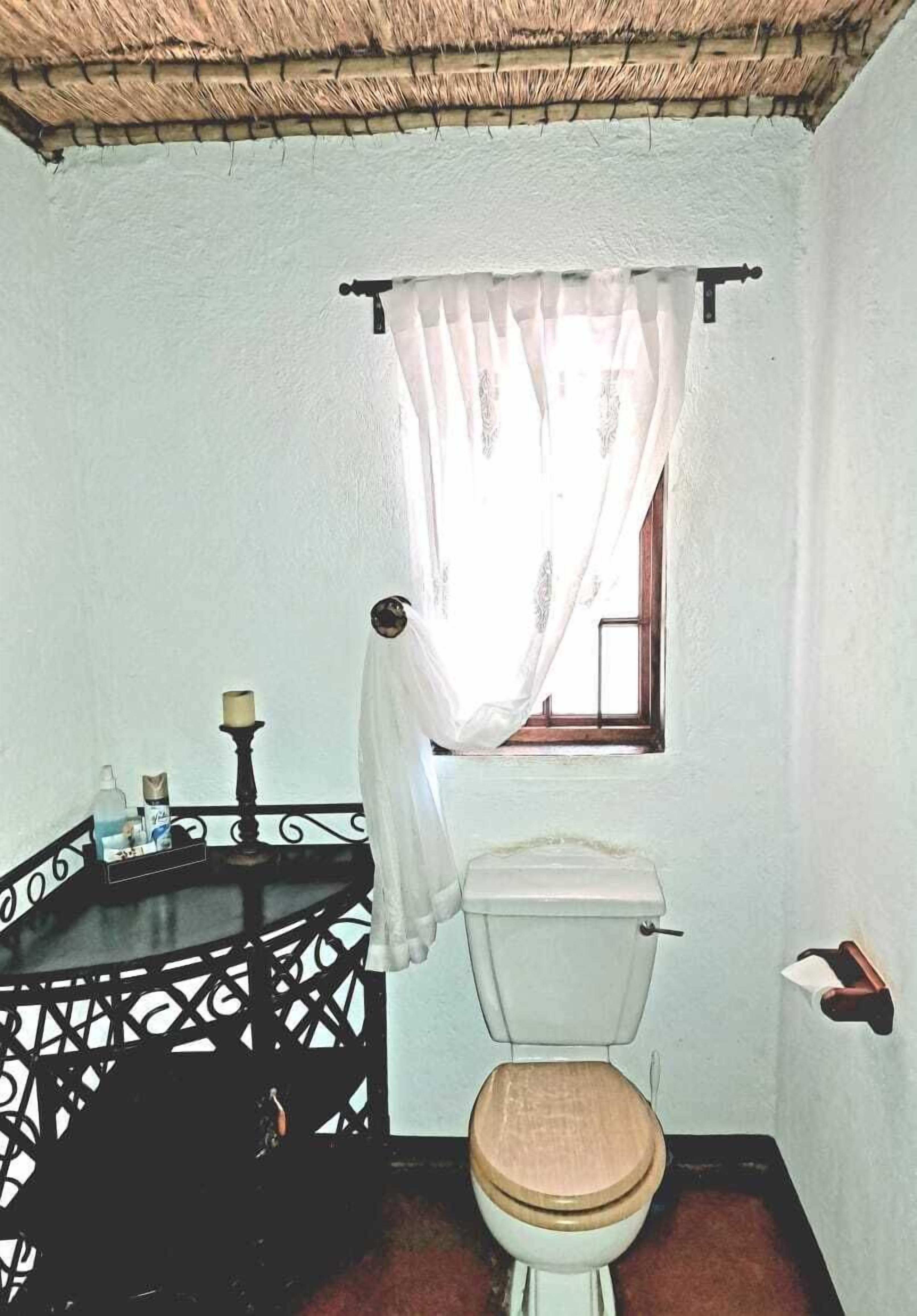 A small bathroom with a toilet, a sink with a black ornate cabinet, a window with a white curtain, and a small shelf with toiletries.