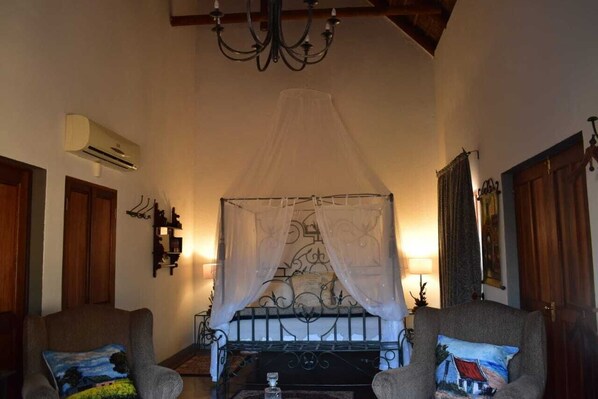 A room with a four-poster bed, two armchairs, a chandelier, an air conditioning unit, and a wall-mounted shelf.