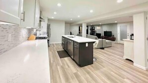 Cozinha moderna com armários brancos, ilha central e área de refeições com sofá e televisão.