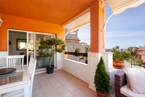 Un balcón con puerta de vidrio, plantas en macetas, un sofá de mimbre y una mesa de comedor.