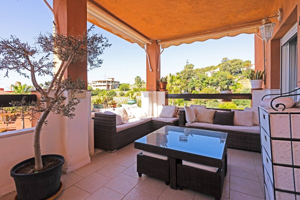 An outdoor patio with wicker furniture, a glass-top table, and a potted tree.