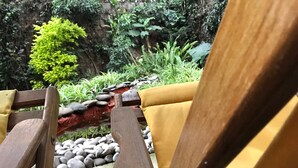 Ein hölzerner Stuhl mit gelber Polsterung, neben einem Garten mit einem steingefassten Wasserspiel und üppigem Grün.