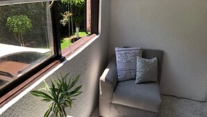 Ein kleiner, moderner Raum mit einem grauen Sofa, einem gemusterten Kissen, einer Topfpflanze und Blick auf einen Garten durch ein Fenster.