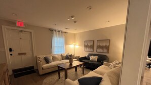 A living room with a beige sofa, a black leather sofa, a coffee table, and a lamp. There is a door with an exit sign, a window with curtains, and two framed pictures on the wall.