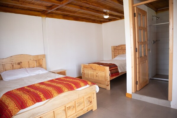 Una habitación con dos camas de madera, una puerta de madera que da a un área de ducha y piso cubierto de baldosas blancas.