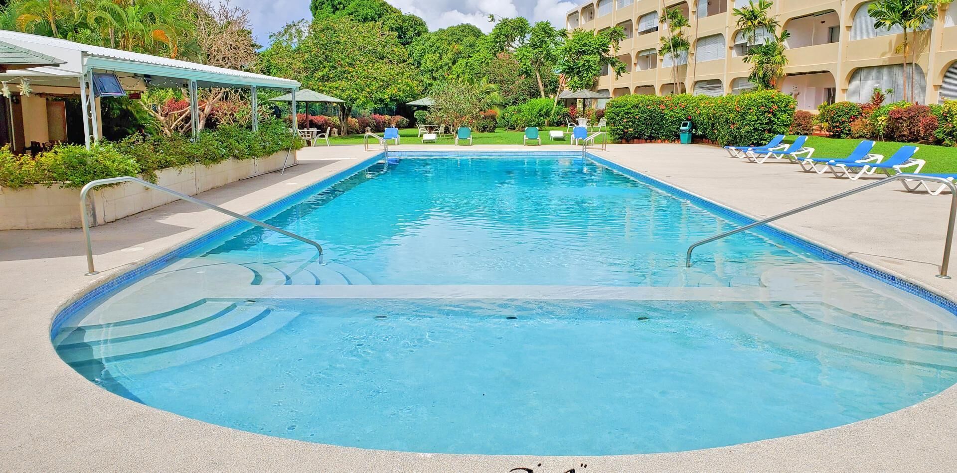 A rectangular swimming pool with a clear blue water surface, surrounded by a concrete deck. There are lounge chairs and umbrellas along the poolside, and a building with multiple balconies in the background.