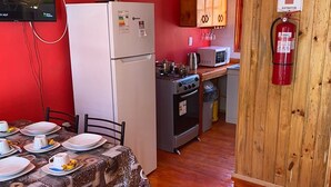 A compact kitchen with a refrigerator, stove, microwave, and wooden cabinets. A dining table with plates and a patterned tablecloth is set up. - Parcela Los Nonos (Vicuña)