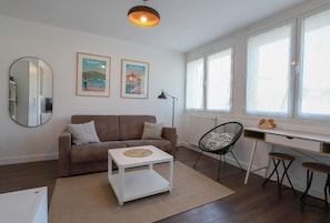 A modern living room with a brown sofa, a white coffee table, a black chair, a desk with stools, and large windows. - Home Sweet Home (Annecy)