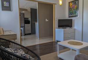 Una sala de estar moderna con un sofá, un televisor sobre un mueble, una mesa de comedor y una cocina al fondo.