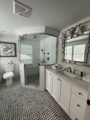 A modern bathroom with a glass-enclosed shower, white tiled walls, a large mirror, and a patterned floor.