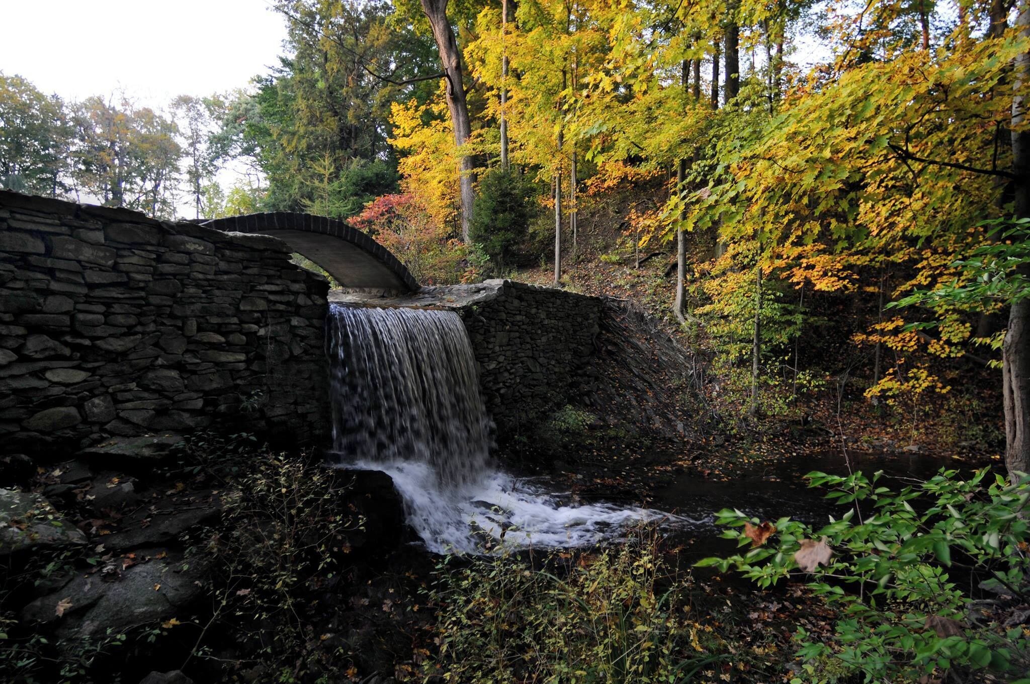 Een stenen brug over een waterval in een bebost gebied met herfstkleuren.