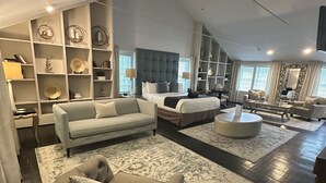 A spacious living room with a large sofa, a round ottoman, and a coffee table. There are shelves with decorative items and a bed with a tufted headboard. - Buttermilk Falls Inn (Milton)