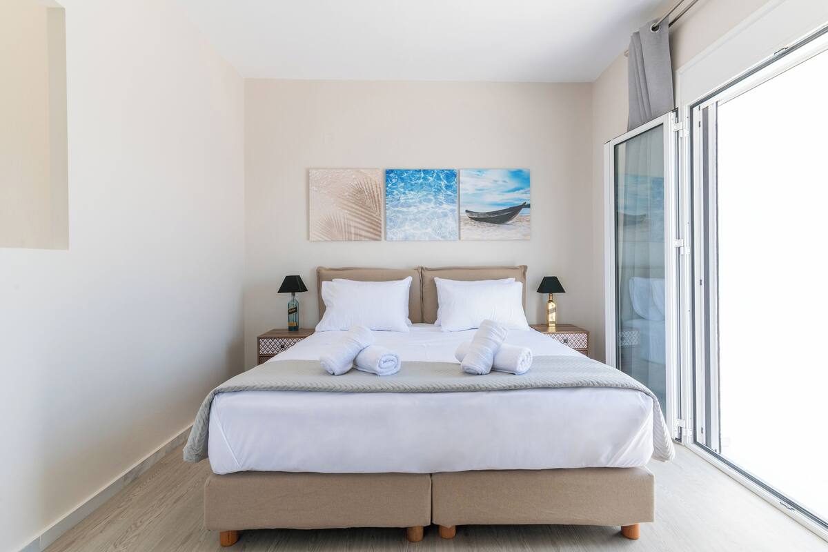 A neatly made bed with white linens, two bedside lamps, and a view of the sea through a sliding glass door.