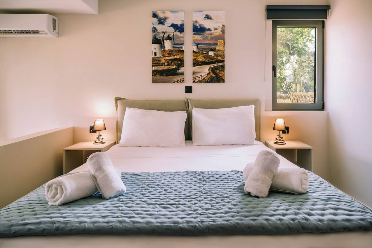A neatly made bed with white linens and two rolled towels, a wall-mounted air conditioner, and two bedside lamps on either side.
