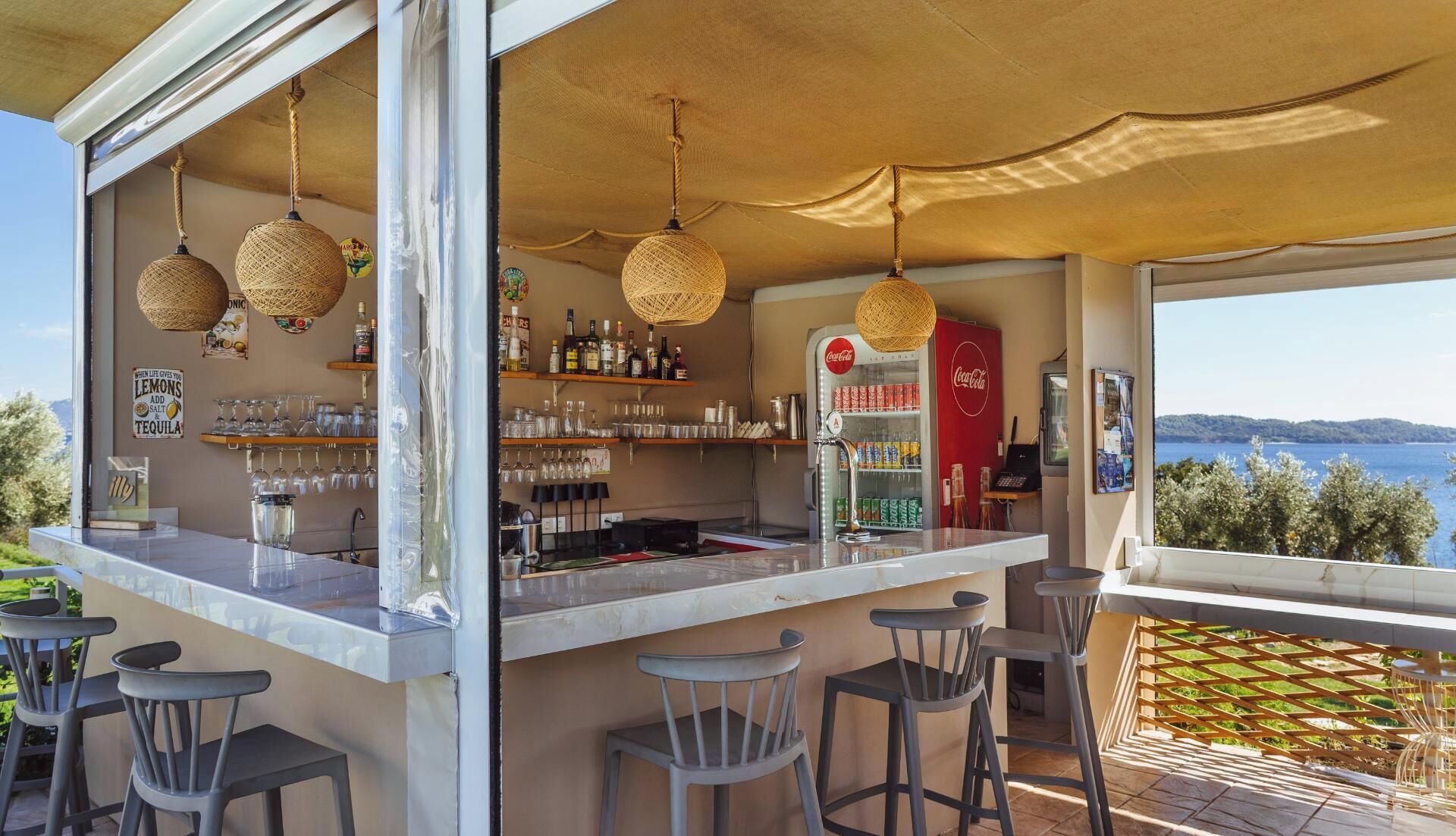 A bar area with a view of the sea, bar stools, and a Coca-Cola refrigerator.