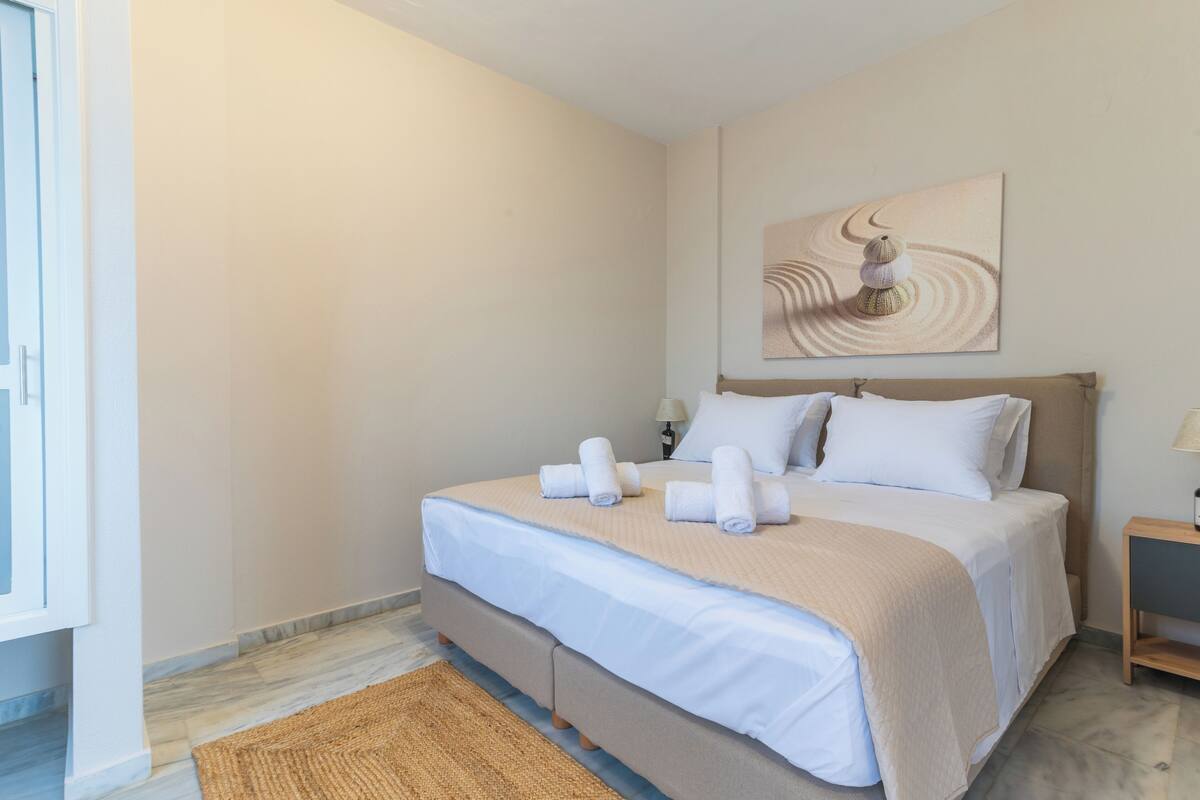 A neatly made bed with white linens and a beige throw blanket, a wooden nightstand with a lamp, and a painting on the wall.