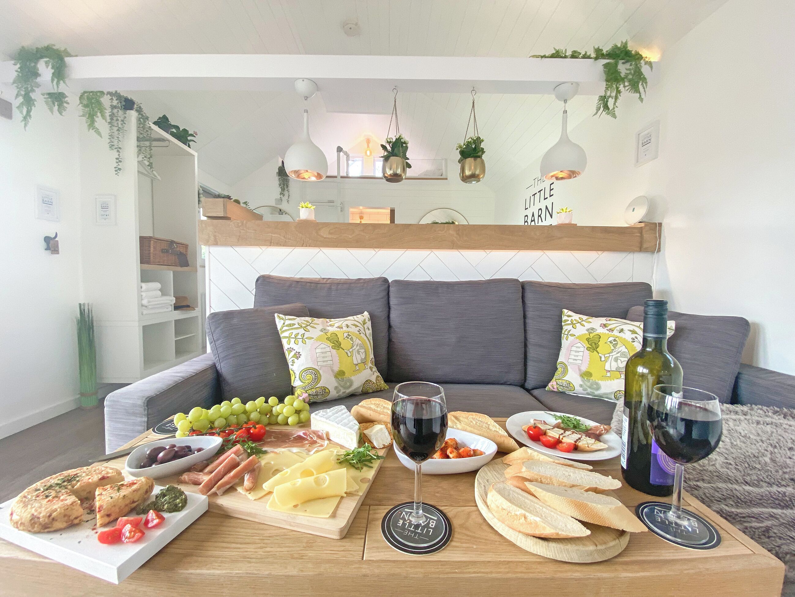 Een gezellige woonkamer met een grijze bank, een houten salontafel met een kaasplank, wijnglazen en een fles wijn.