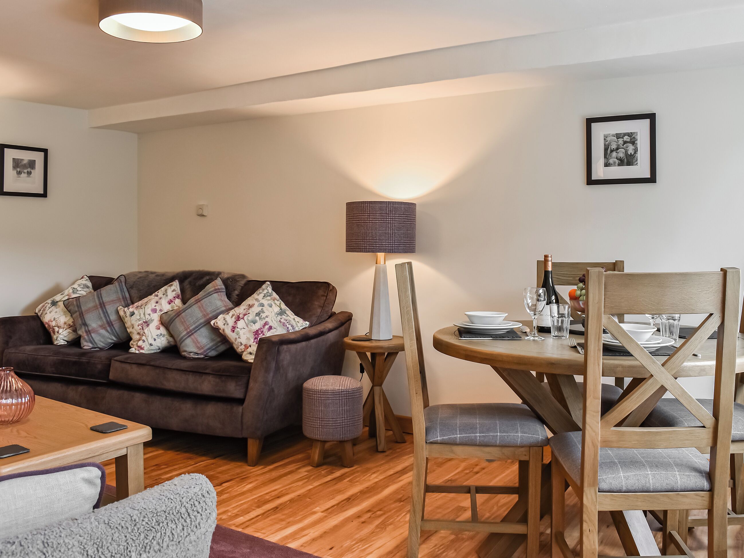 A cozy living room with a brown sofa, a wooden dining table set for two, and a small side table with a lamp.