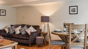 Un salon chaleureux avec un canapé marron, une table à manger en bois dressée pour deux personnes et une petite table d’appoint avec une lampe.
