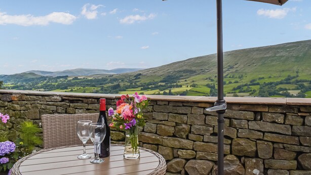 Une table avec une bouteille de vin, deux verres et un vase de fleurs, le tout placé devant un mur de pierre, avec une vue panoramique sur des collines verdoyantes et un ciel dégagé.