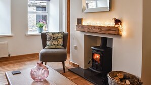 Un salon chaleureux avec une cheminée, un fauteuil agrémenté d’un coussin à motifs, une table en bois sur laquelle est posé un vase, et un panier rempli de bûches.