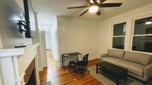 Un salon avec une cheminée, un canapé, une table basse, un bureau avec une chaise et un ventilateur de plafond.