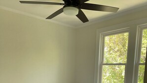 Une chambre à coucher avec un lit, un ventilateur de plafond, une table de chevet avec une lampe et une fenêtre donnant sur des arbres.