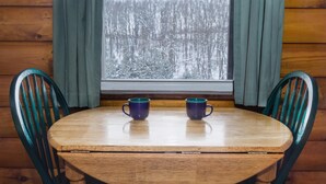 A wooden table with two blue cups, surrounded by green chairs, in a cabin with a window showing a snowy forest. - Dove Cottage at Secluded Cabins (Lakeville)