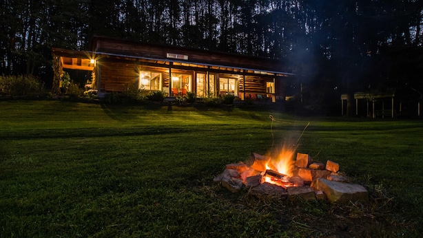A cabin with a fire pit in the foreground, surrounded by a well-maintained lawn and a forest in the background.