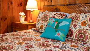 Una cama con ropa de cama floral, un cojín color turquesa con un diseño de pájaro, un cabecero de madera y una mesita de noche con una lámpara y un pequeño jarrón de flores.