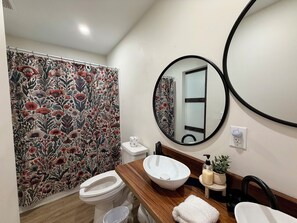 A bathroom with a floral shower curtain, a round mirror, a wooden vanity, a white sink, and a toilet. - 6BR Crew Stay - Kitchen - Parking - Near I-26 - C (North Charleston)