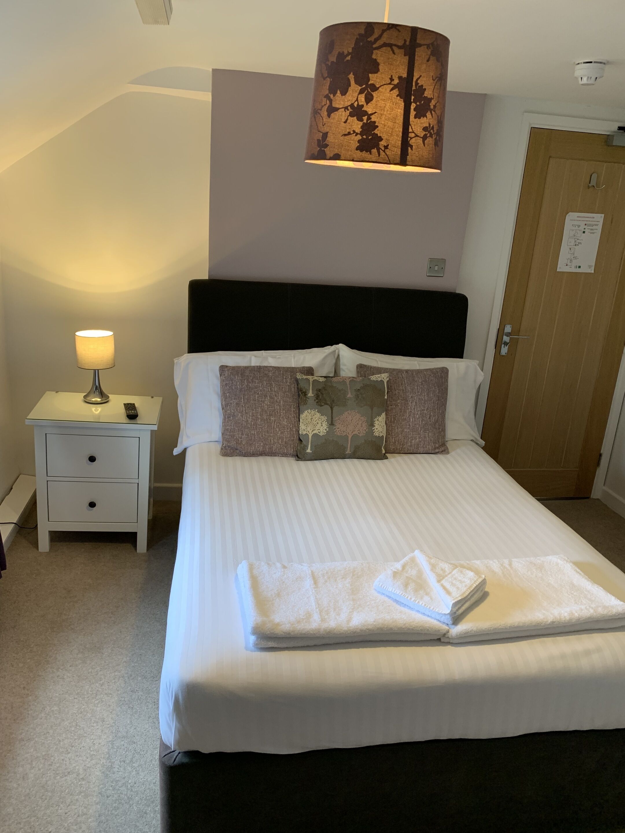A neatly made bed with a white comforter and pillows, a bedside lamp, a small bedside table with a remote control, and a door with a sign on it.
