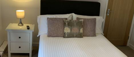 A neatly made bed with a white comforter and pillows, a bedside lamp, a small bedside table with a remote control, and a door with a sign on it.