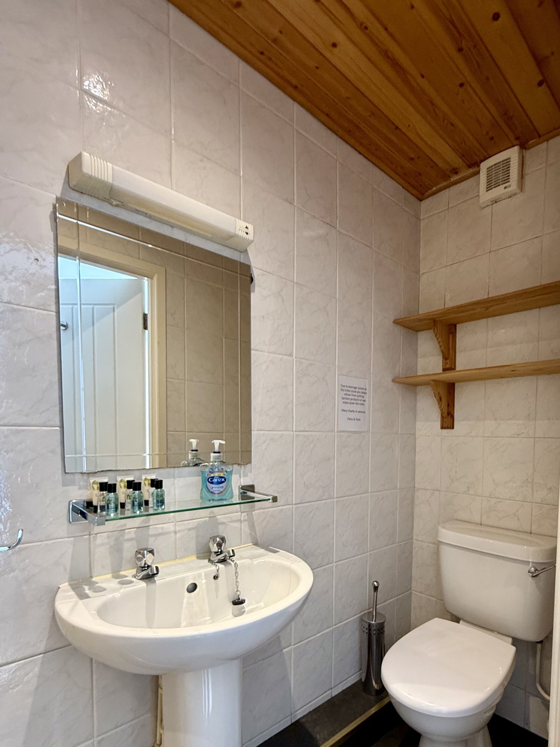 A bathroom with a white sink, mirror, and toilet. There are shelves above the sink and a vent on the wall.