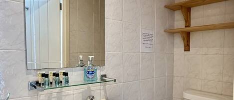 A bathroom with a white sink, mirror, and toilet. There are shelves above the sink and a vent on the wall.