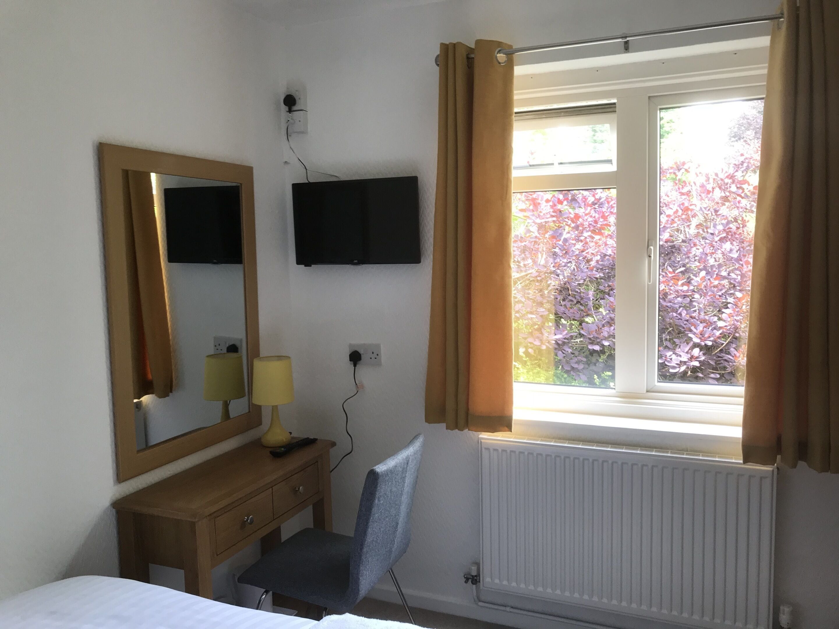 A hotel room with a bed, a desk with a chair, a mirror, a flat-screen TV mounted on the wall, and a window with curtains.