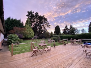 Una terraza de madera con mobiliario exterior, rodeada de un césped cuidado y árboles, bajo un cielo crepuscular.