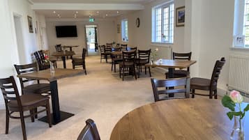 A spacious dining area with multiple wooden tables and chairs, a flat-screen TV mounted on the wall, and large windows allowing natural light.