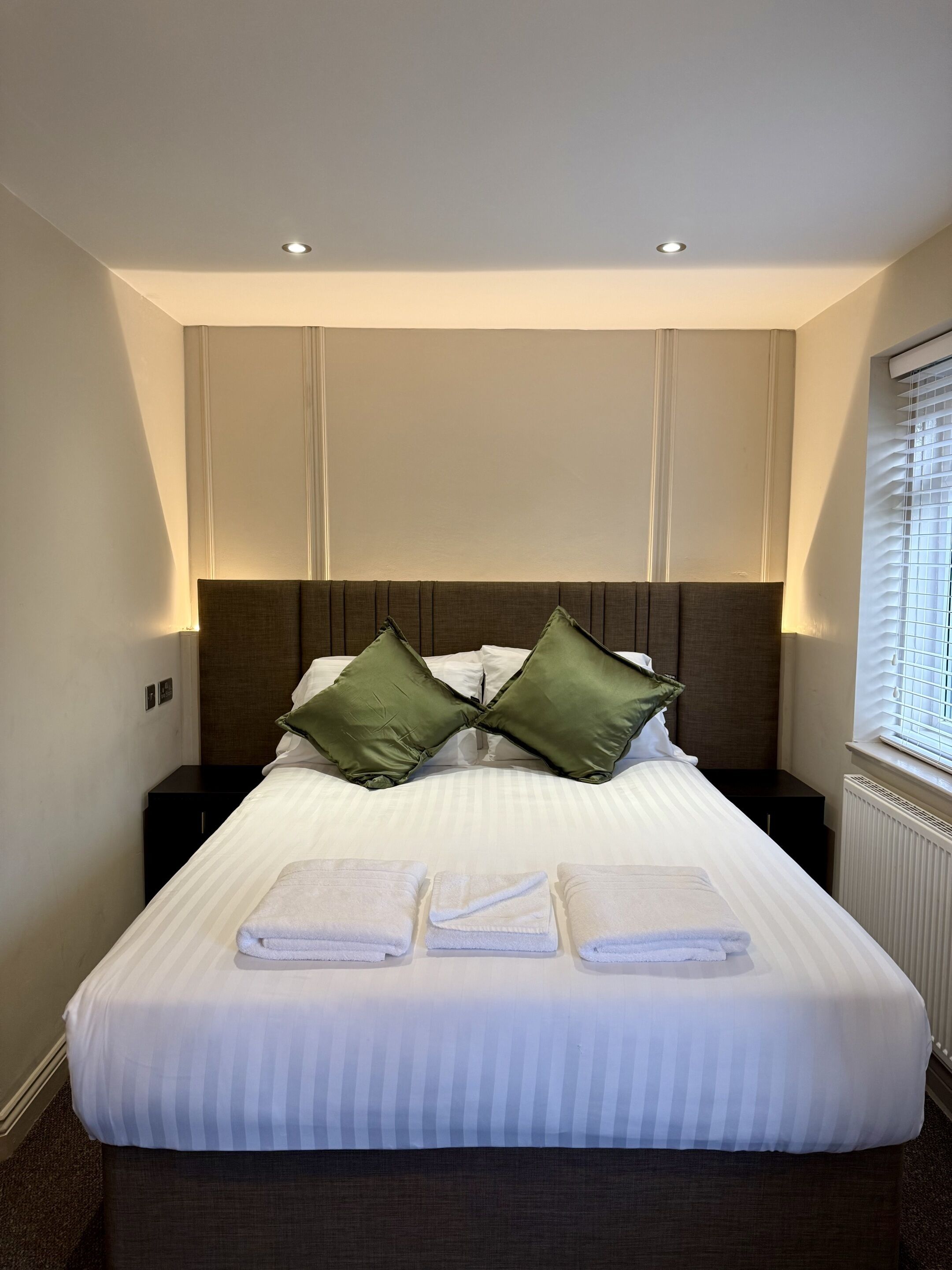 A neatly made bed with white linens and two green pillows, a dark headboard, and a window with blinds on the right.