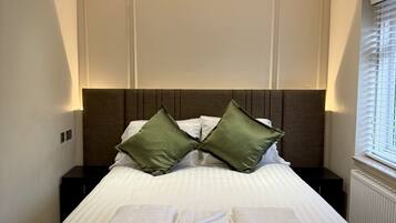 A neatly made bed with white linens and two green pillows, a dark headboard, and a window with blinds on the right.