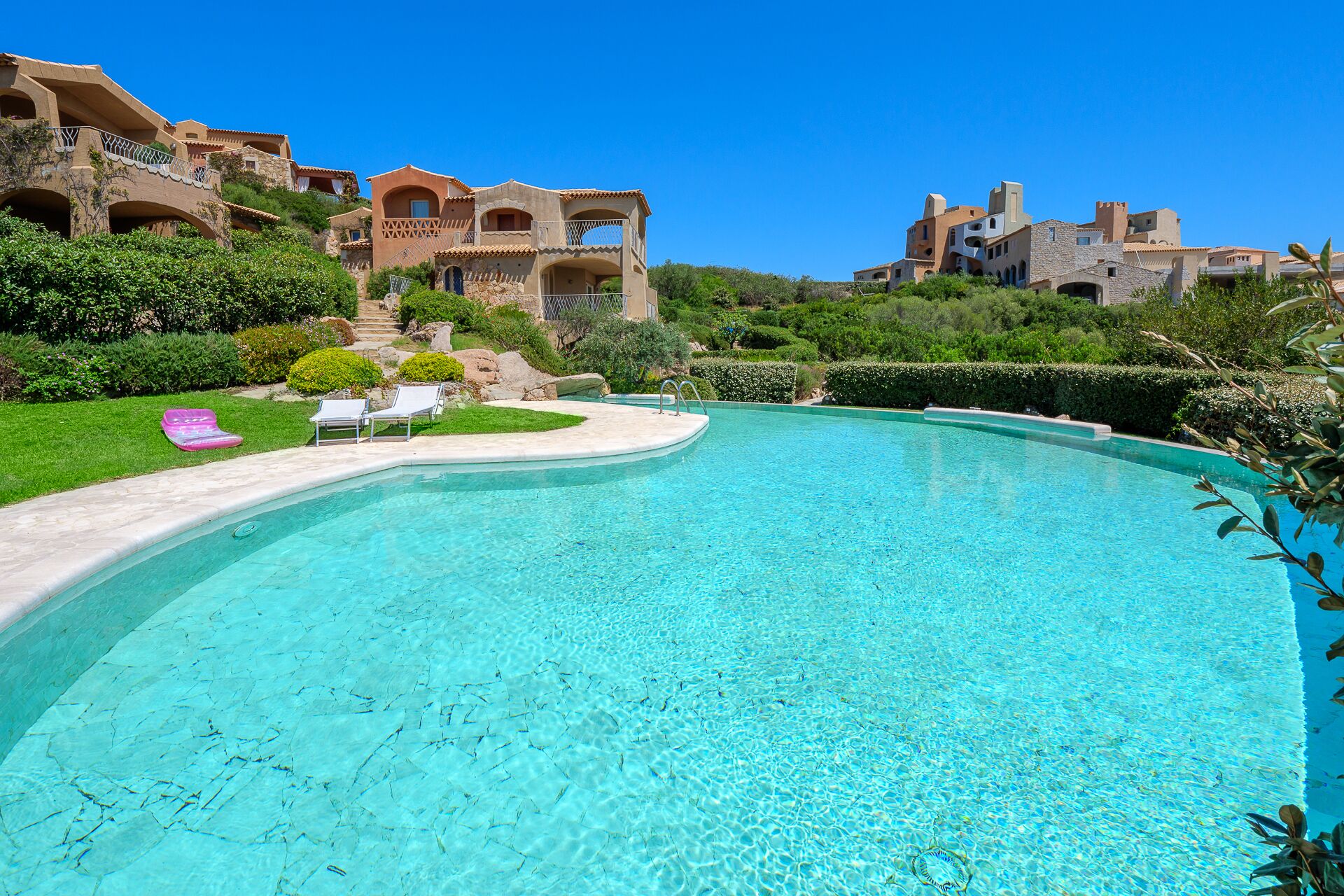 A clear blue pool with lounge chairs and a stone bridge.