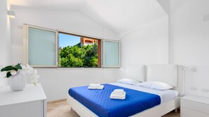 Ein modernes Hotelzimmer mit Bett, weißen Nachttischen, einem Schreibtisch und einem Fenster mit Blick auf Grünflächen und ein Gebäude.
