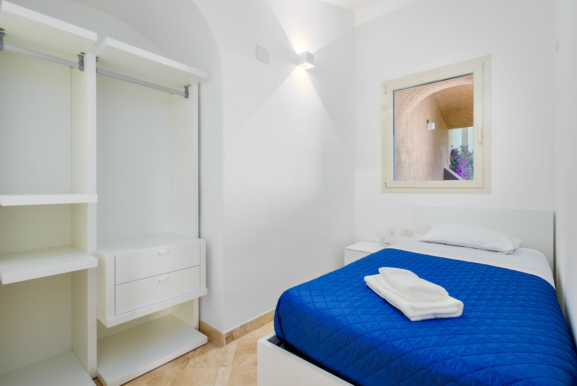 A bedroom with a bed, a nightstand, a wardrobe, and a window with a view of an outdoor area.