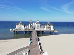 Une jetée mène à un bâtiment blanc doté de nombreuses fenêtres et d’une entrée centrale, qui surplombe une plage et l’océan.