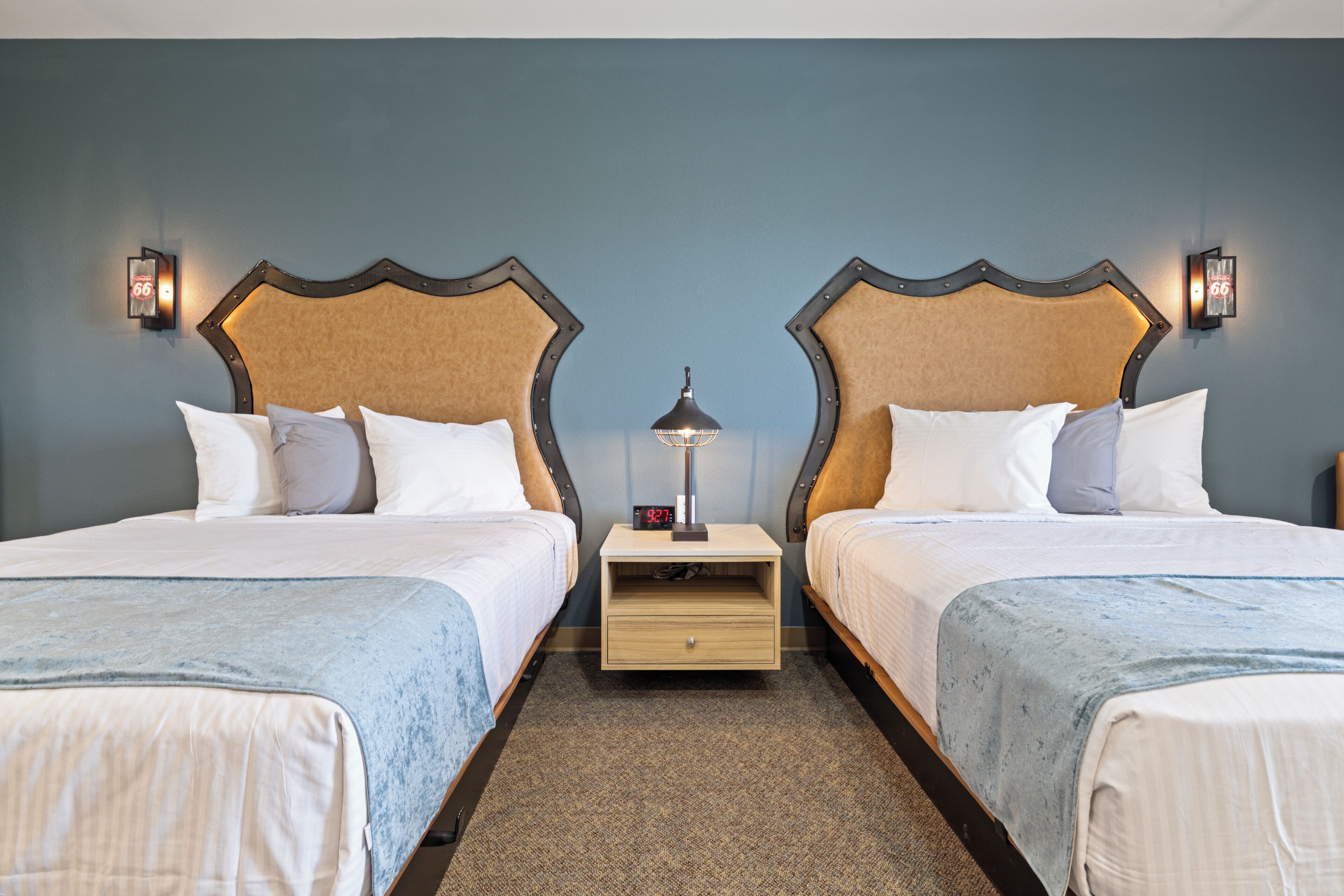 A hotel room with two beds, each with a headboard, a nightstand with a lamp, and a digital clock.