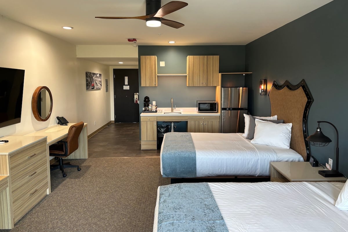 A hotel room with a desk, two beds, a kitchenette, and a ceiling fan.