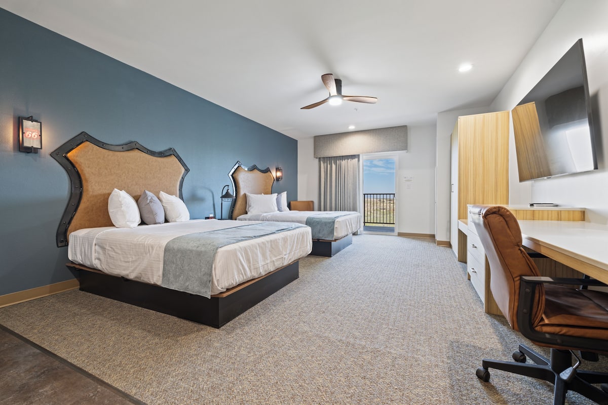 A modern hotel room with two beds, a desk, a chair, a TV, and a balcony with a view.