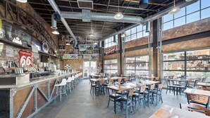 A spacious restaurant with high ceilings, exposed ductwork, and large windows offering views of the city. Tables and chairs are neatly arranged, and a bar area is visible on the left. - Hotel Topock66 (Topock)