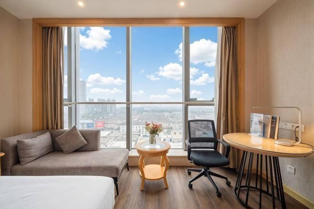 A hotel room with a large window, a bed, a sofa, a small round table, and a chair.